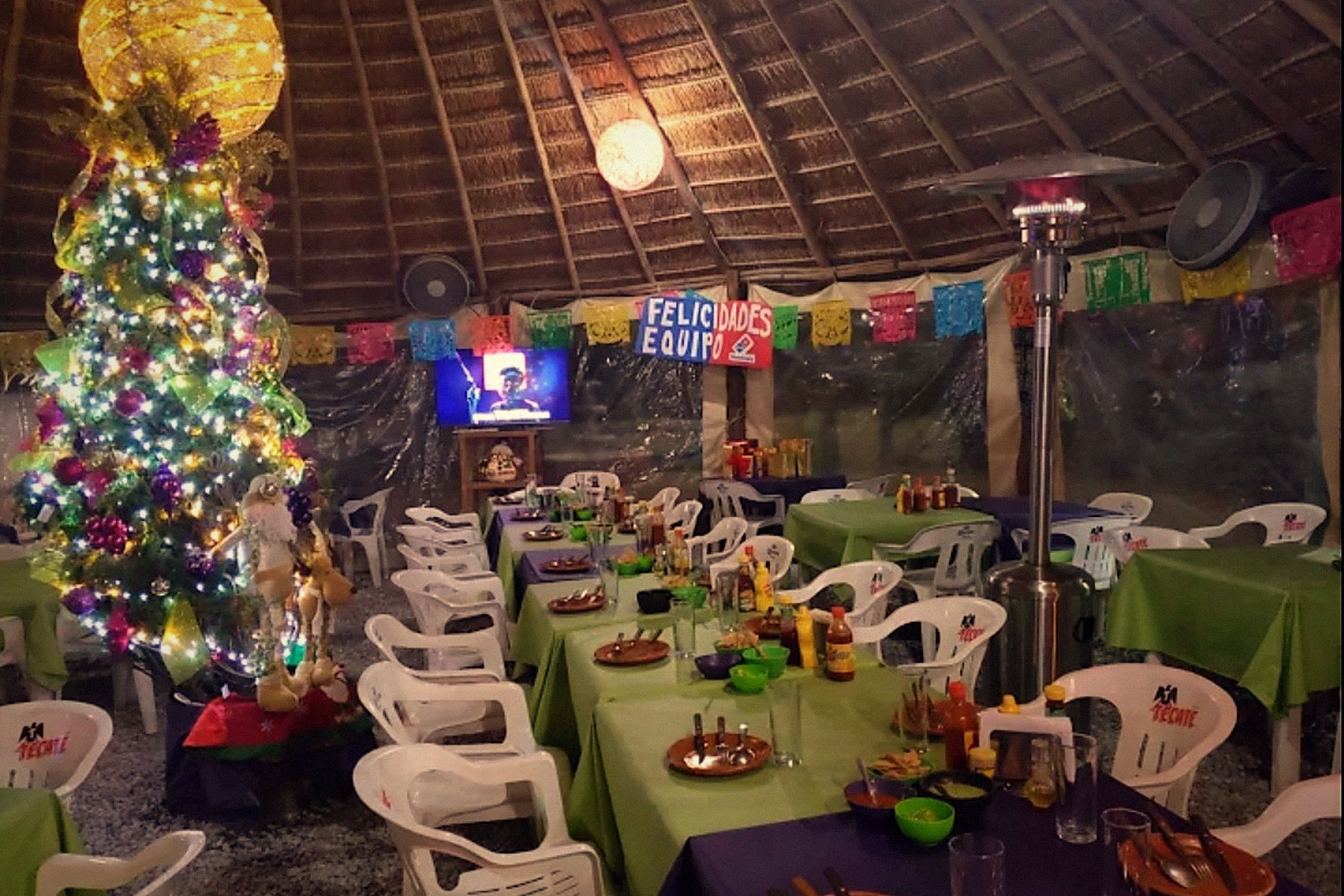 Mesas largas servidas de noche bajo la palapa, con papel picado y calentadores
